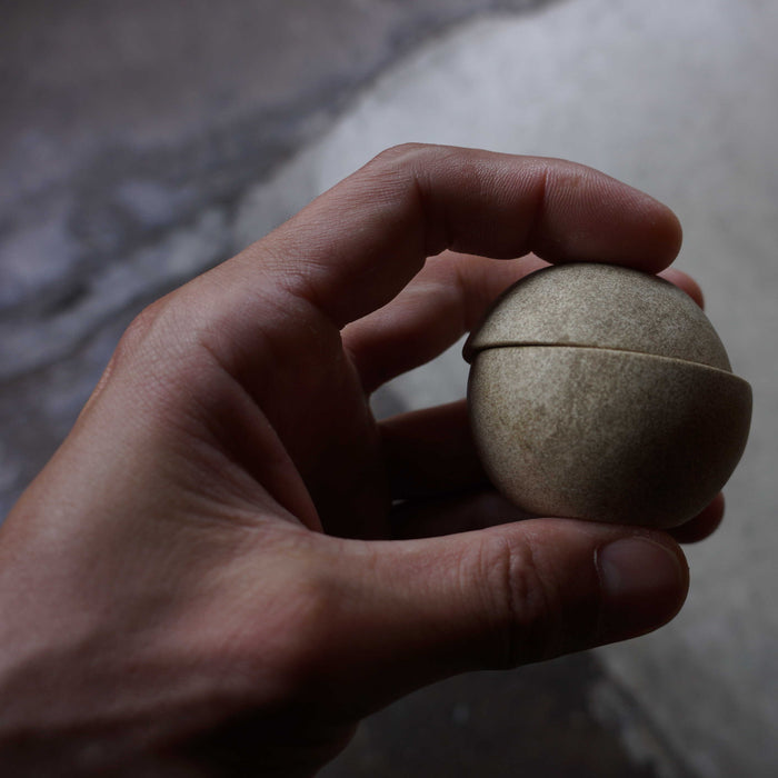 Hand holding a minimalist spherical candle holder in a light stone finish, shown close up against a moody neutral background to highlight its compact size and sculptural design.