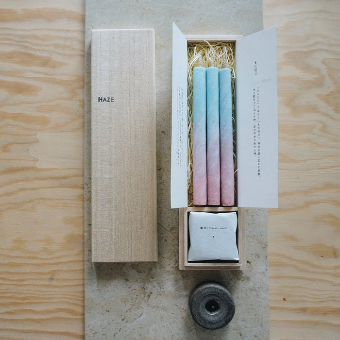 Gift set of three pastel blue and pink taper candles in a wooden HAZE box with candle stand and packaging inserts, displayed on a neutral stone and wood surface in a minimalist product presentation.