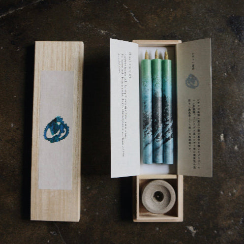Gift set of three blue and green marbled taper candles in a wooden presentation box with candle holder and printed inserts, displayed on a dark textured surface in a minimalist art-inspired product presentation.