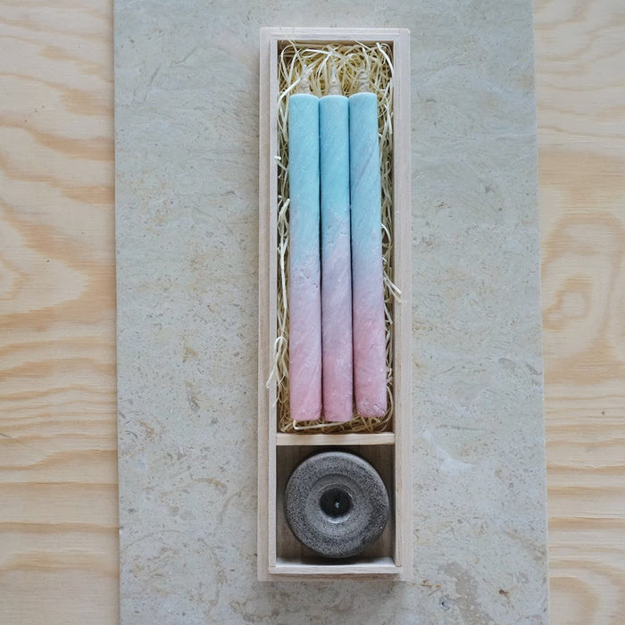 Gift set of three pastel blue and pink taper candles with a stone candle holder, arranged in a wooden presentation box on a neutral stone and wood surface in a minimalist product display.