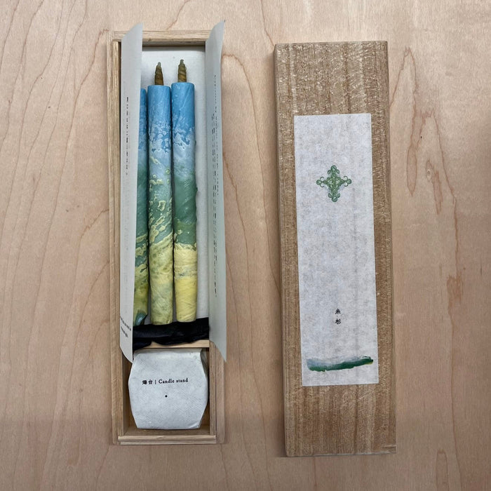 Gift set of two blue and green marbled taper candles with a candle stand in a wooden presentation box, displayed on a light wood surface in a minimalist art-inspired product presentation.