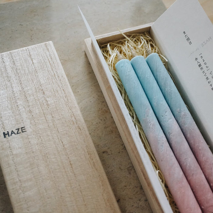 Close-up of a HAZE wooden gift box containing three pastel blue and pink taper candles with packaging inserts, styled on a neutral stone surface in a minimalist product presentation.