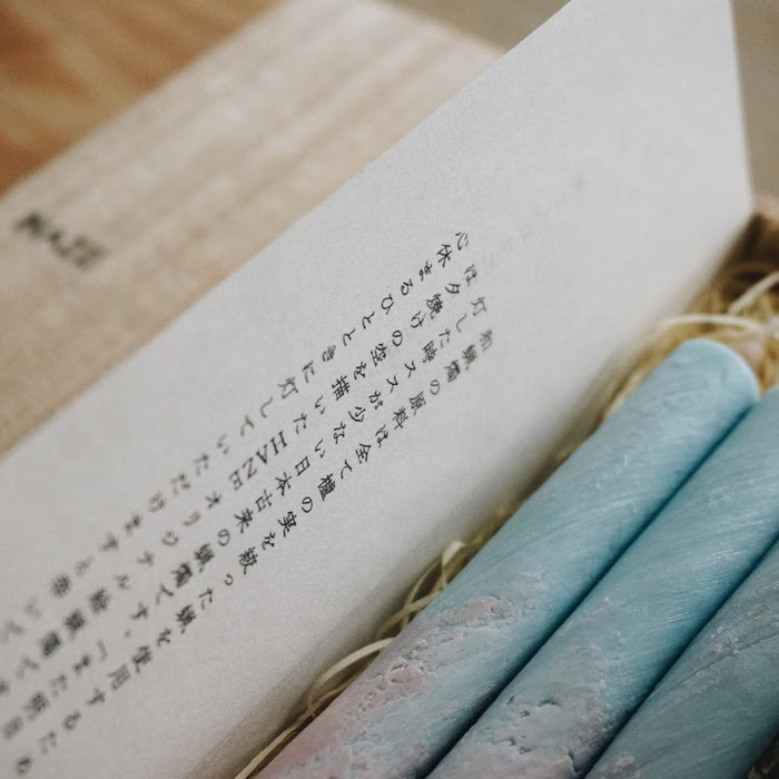 Close-up of pastel blue and pink taper candles inside a HAZE wooden gift box with printed insert card, styled in a minimalist art-inspired product presentation.