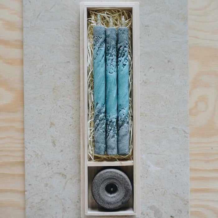 Gift set of three blue, gray, and white marbled taper candles with a stone candle holder, arranged in a wooden presentation box on a neutral stone and wood surface in a minimalist art-inspired product display.