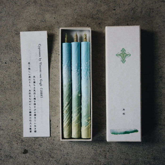 Set of three art-inspired blue and green taper candles in an open presentation box with printed insert card, styled on a concrete surface in a minimalist product display.