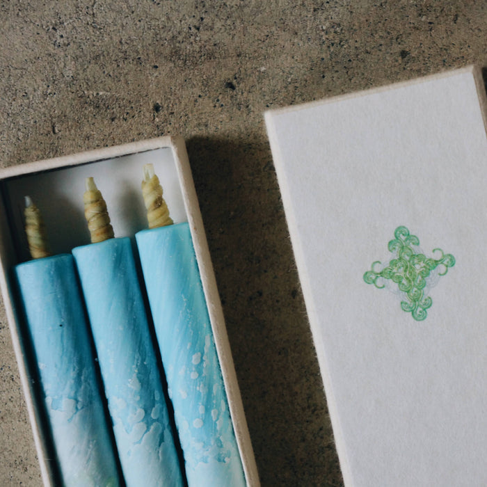 Close-up of three blue art-inspired taper candles in a white presentation box with decorative sleeve, styled on a concrete surface in a minimalist product display.