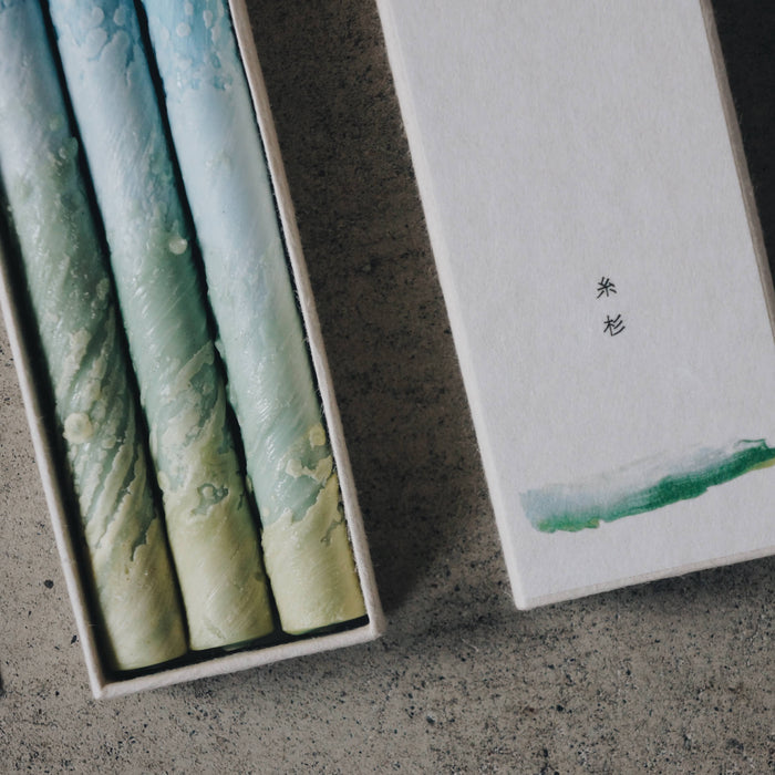 Close-up of three blue and green marbled taper candles in a minimalist presentation box with decorative sleeve, styled on a concrete surface in an art-inspired product display.