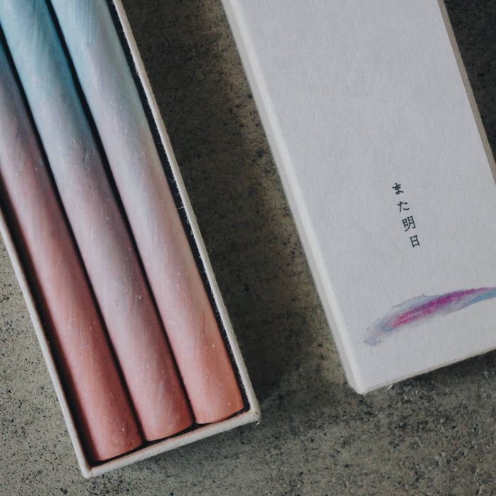 Close-up of three pastel blue and pink taper candles in a minimalist presentation box with decorative sleeve, styled on a concrete surface in an art-inspired product display.