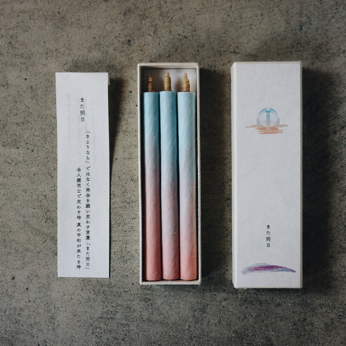 Set of three pastel blue and pink taper candles in an open presentation box with printed insert card and decorative sleeve, styled on a concrete surface in a minimalist art-inspired product display.