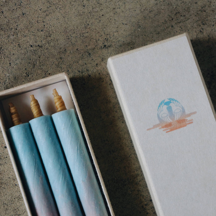 Close-up of three pastel blue and pink taper candles in a white presentation box with matching decorative sleeve, styled on a concrete surface in a minimalist art-inspired product display.