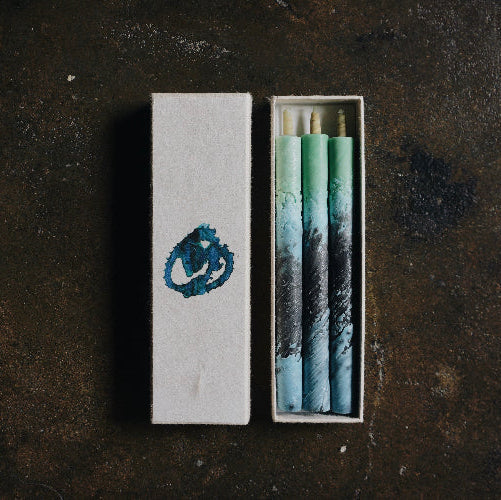 Set of three blue and green marbled taper candles in an open presentation box with matching decorative sleeve, styled on a dark textured surface in a minimalist art-inspired product display.