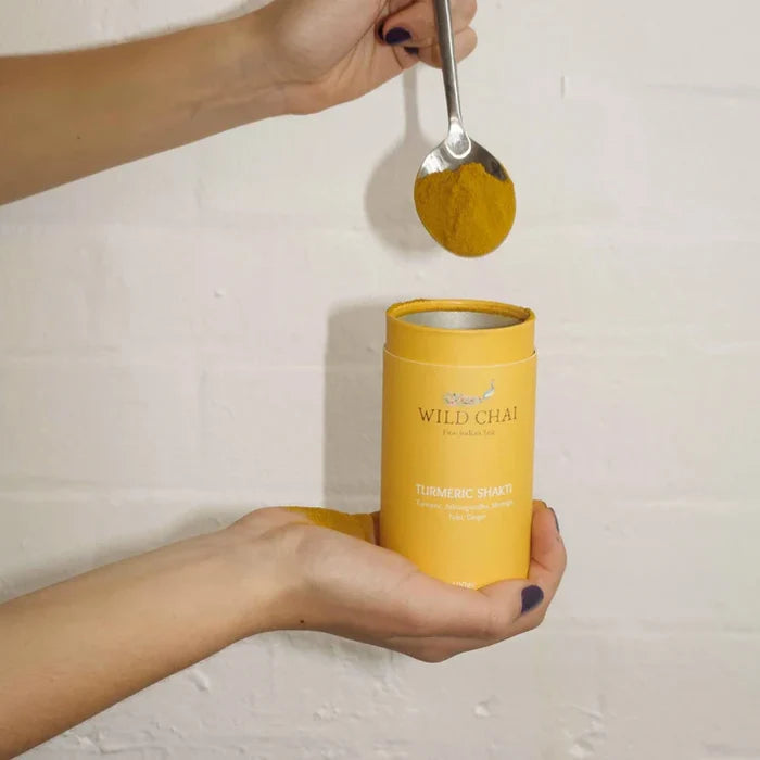 Person holding a yellow 'Wild Chai' container with a spoonful of spice above it against a white background.